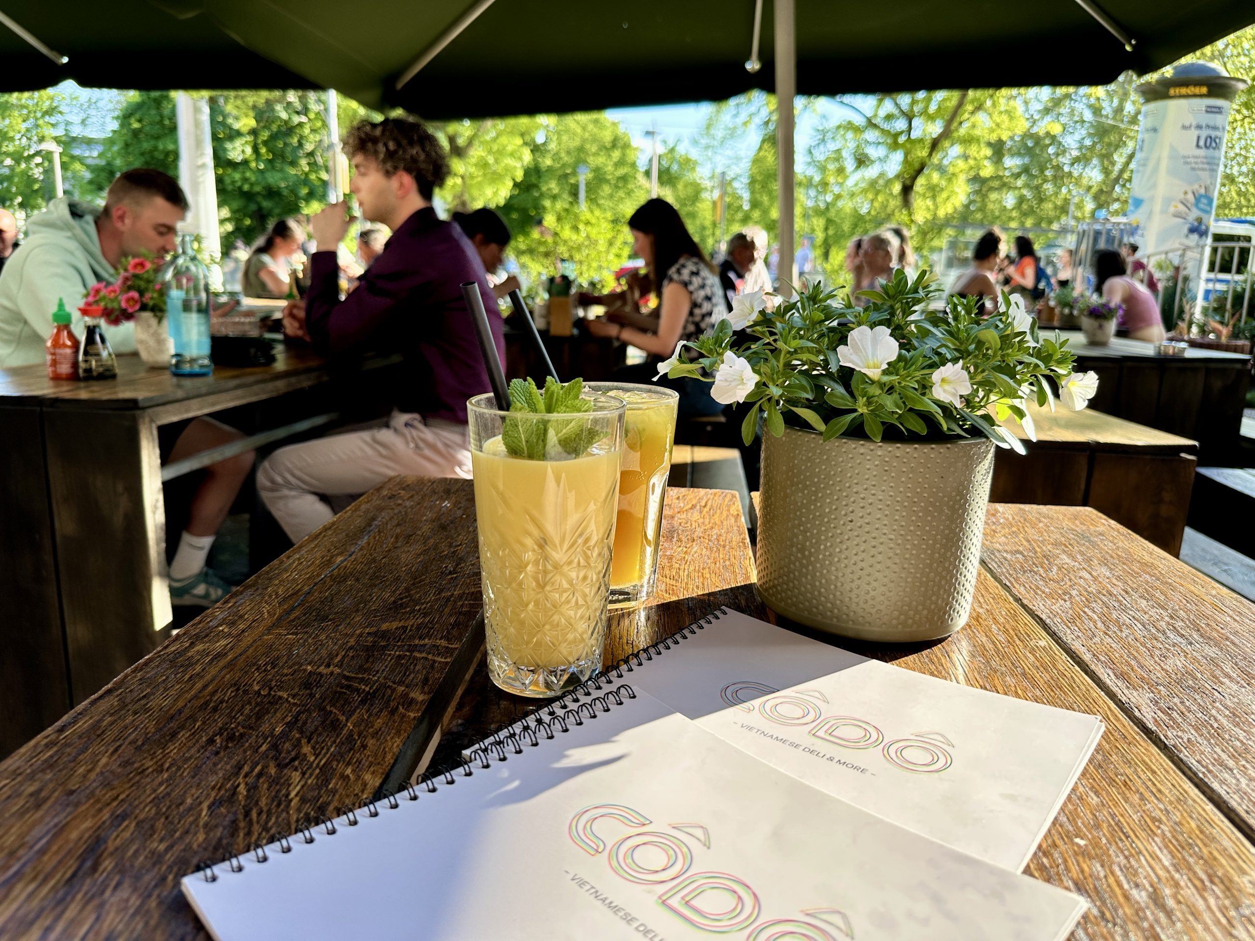Zwei helle Drinks mit Minze auf einem Holztisch neben CÔDÔ-Menükarten und einem Blumentopf; Gäste sitzen im Hintergrund auf der Terrasse.