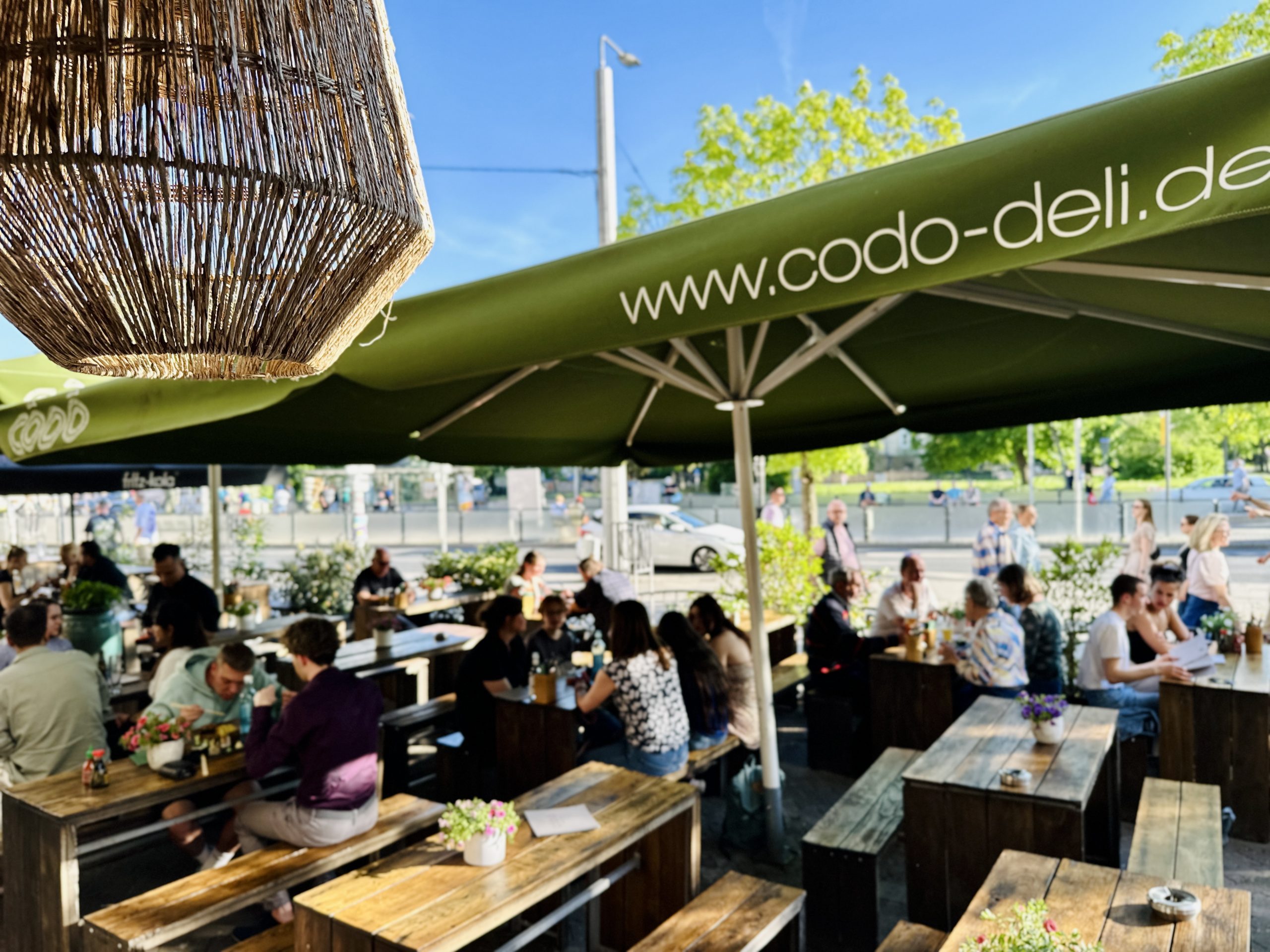 Außenbereich mit Gästen an Holztischen unter großen Sonnenschirmen mit Aufdruck „www.codo-deli.de“ an einem sonnigen Tag.