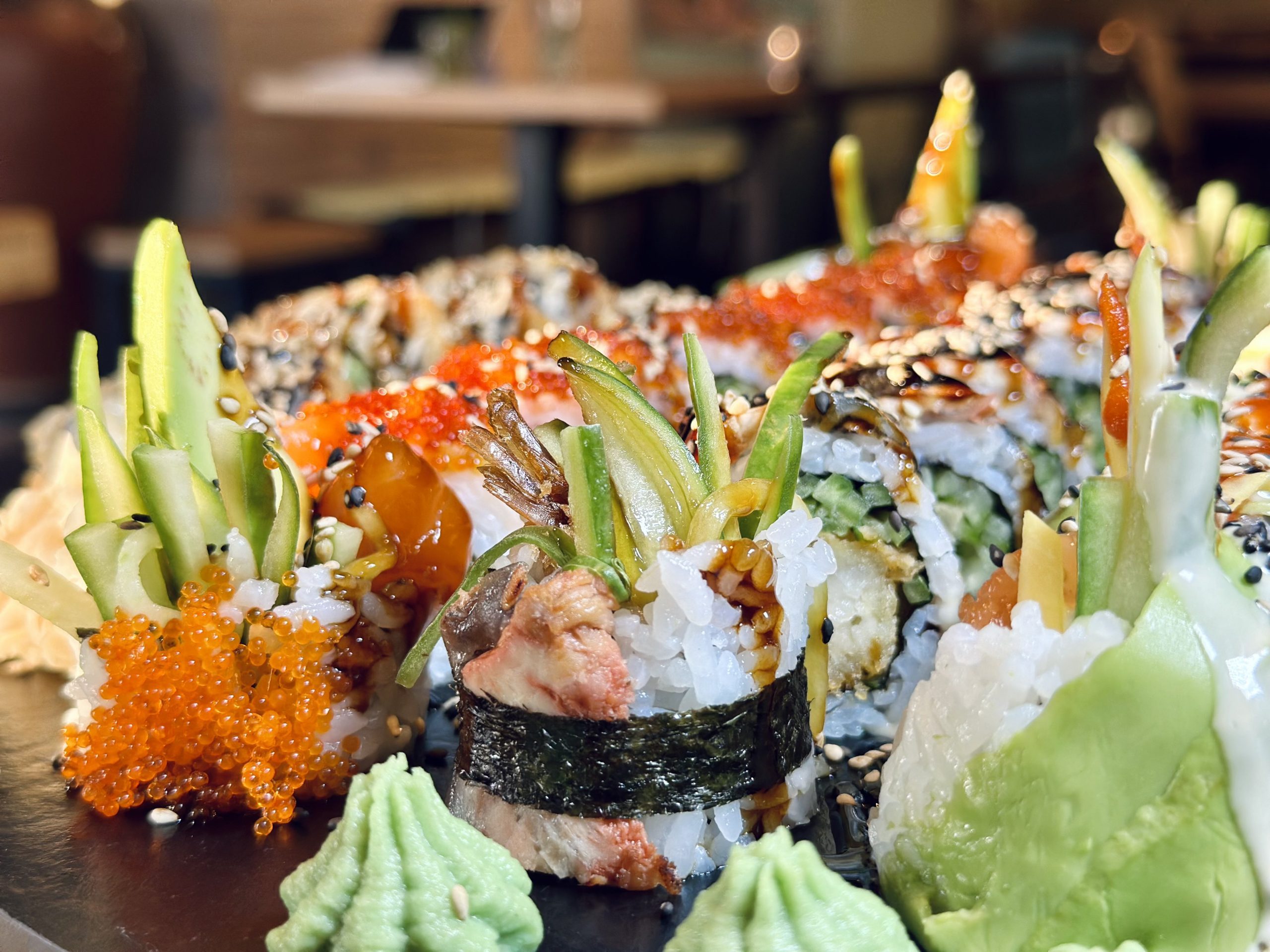 Nahaufnahme von Sushi-Rolls mit Avocado, Sesam und Sauce; dazu Wasabi und orangefarbener Fischrogen auf dem Teller.