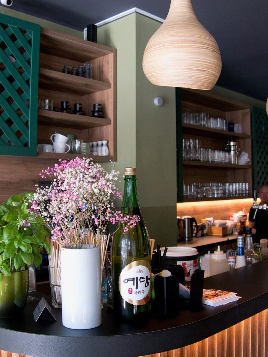 Bar-Theke mit Basilikumtopf, Blumen und großer Flasche; dahinter Holzregale mit Gläsern und warme Hängelampen, Mitarbeitende im Hintergrund.
