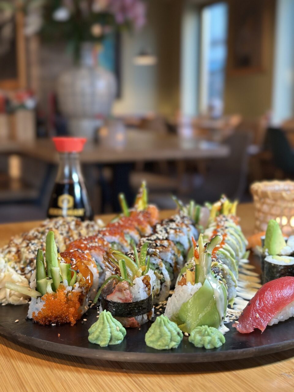 Sushi-Platte mit verschiedenen Rolls, Nigiri, Wasabi und Sojasauce-Flasche im Hintergrund auf einem Restauranttisch.