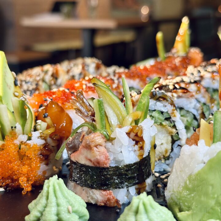 Nahaufnahme von Sushi-Rolls mit Avocado, Sesam und Sauce; dazu Wasabi und orangefarbener Fischrogen auf dem Teller.