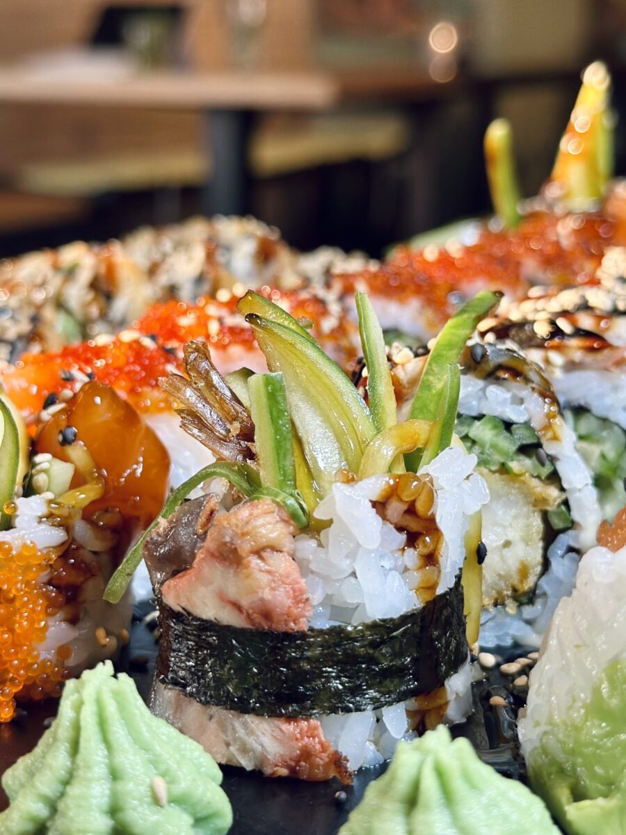 Nahaufnahme von Sushi-Rolls mit Avocado, Sesam und Sauce; dazu Wasabi und orangefarbener Fischrogen auf dem Teller.