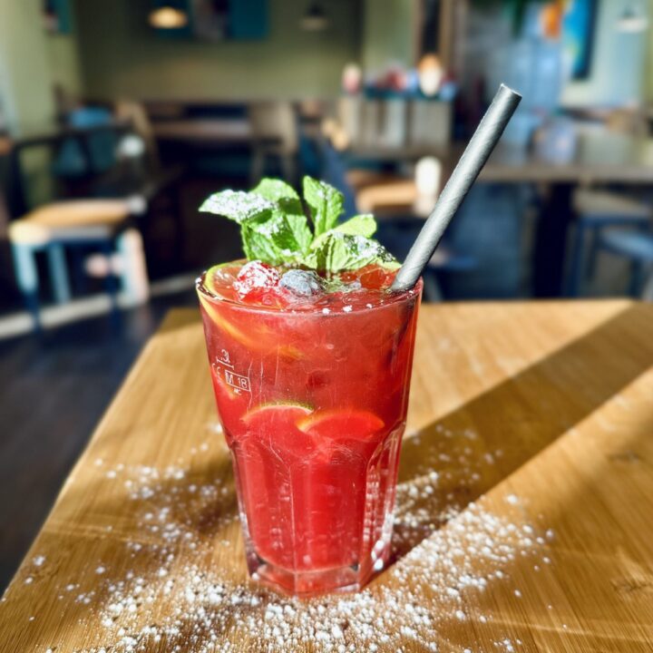 Roter Cocktail mit Eis, Minze und Zitrus-Scheiben im Glas auf einem Holztisch im Restaurant.