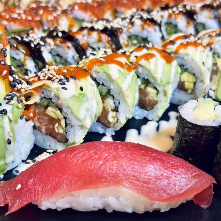 Nahaufnahme von Sushi-Rolls mit Avocado, Sesam und Sauce sowie einem Nigiri im Vordergrund auf einem Teller.