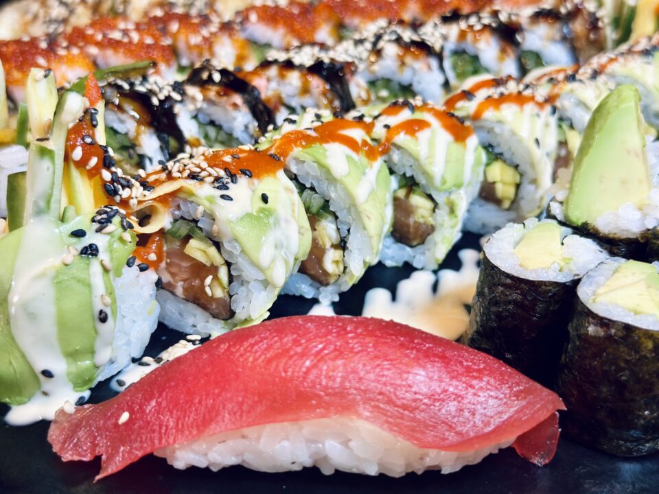 Nahaufnahme von Sushi-Rolls mit Avocado, Sesam und Sauce sowie einem Nigiri im Vordergrund auf einem Teller.